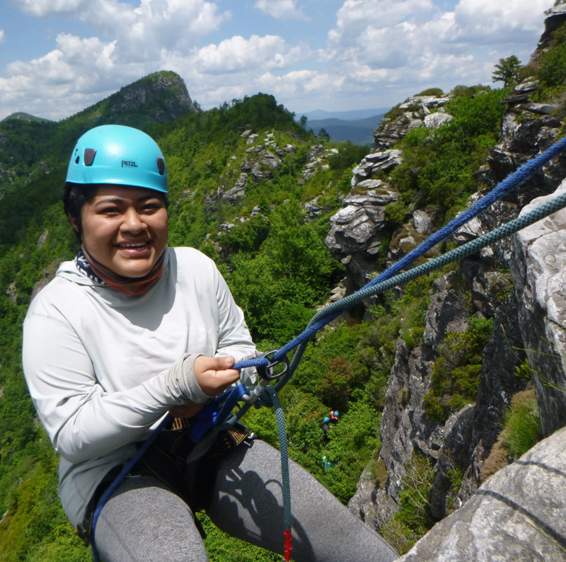 A woman wearing a blue helmet is rappelling down a rocky cliff face. She is holding onto the rope with both hands and smiling at the camera. In the background, there are lush green trees and mountains under a partly cloudy sky. Other people can be seen rappelling further down the cliff.